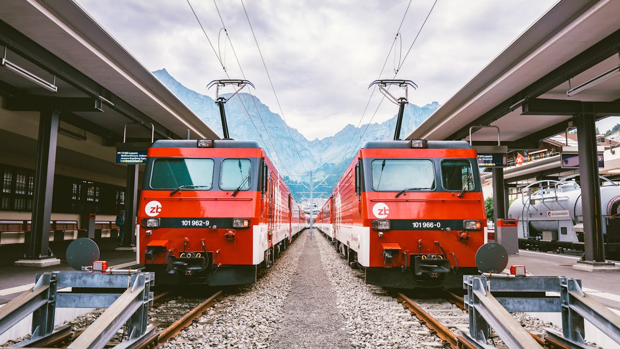 Two red trains at a Swiss station with mountains in the background.